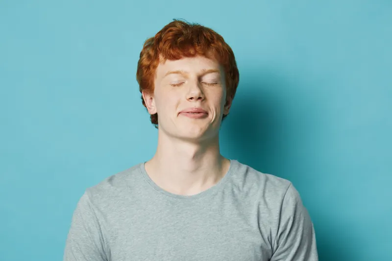 Young man with red hair and closed eyes in gray shirt standing against blue background, calm expression, portrait of relaxed male with light skin and natural hairstyle.