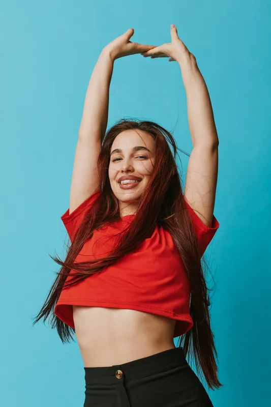 Happy woman with long hair wearing red shirt and black pants stretching arms up against blue background. Smiling girl with closed eyes and relaxed pose in casual outfit indoors.