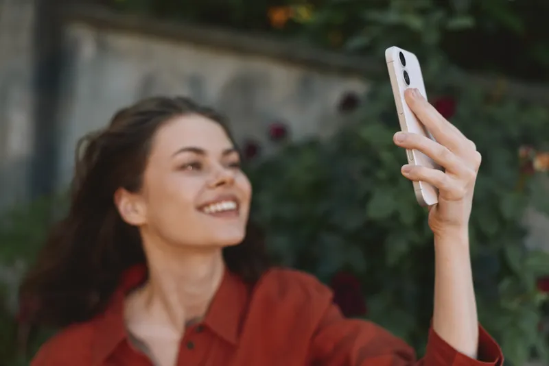 Cheerful woman outdoors, smiling in a sunny selfie moment, red top, relaxed pose, natural colors, soft breeze, lush greenery background, lifestyle vibe, candid