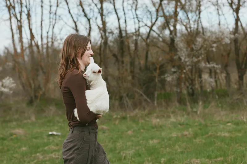 Joyful woman holding a white goat kid outdoors in a park. Natural light and candid lifestyle moment highlighting bond between human and animal, nature, and care.