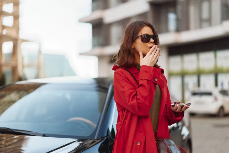 A stylish adult woman in a red coat stands beside a car on a city street, wearing sunglasses and holding a cigarette, exuding confident candid street style in natural urban light