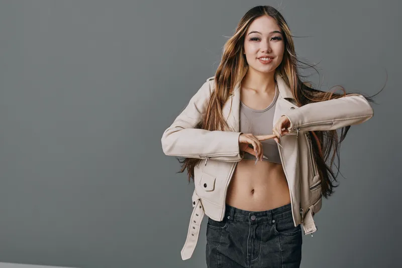 Young woman making heart shape with hands, smiling and posing in leather jacket, casual style and confident expression on gray background.