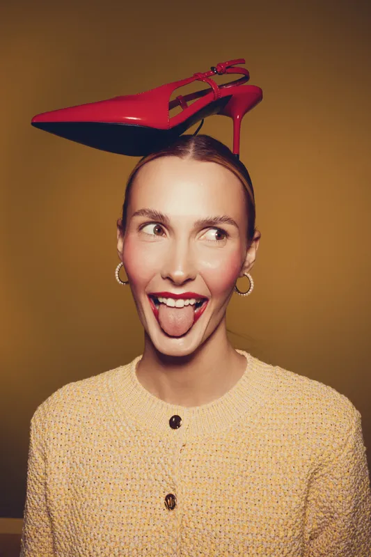 Young woman with red high heel shoe balanced on her head sticks out tongue playfully. She wears hoop earrings and a textured beige sweater against a warm brown background. Concept of fun and fashion.