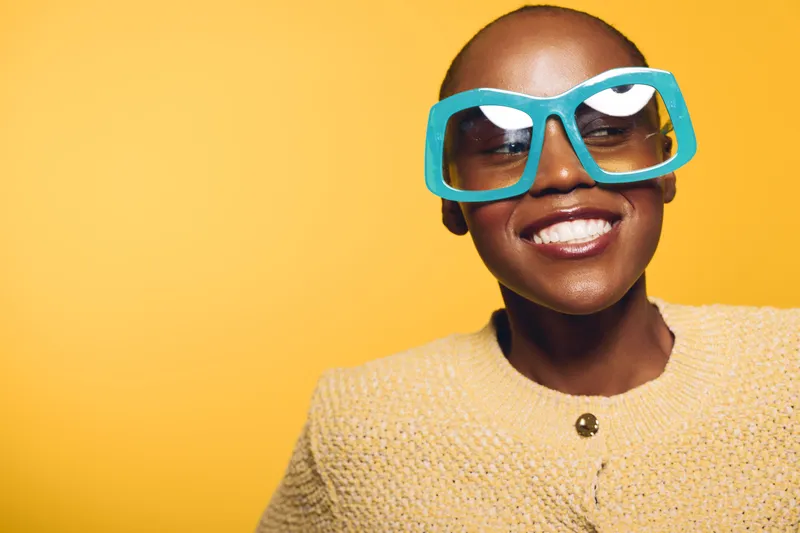 Smiling African American woman with dark skin wearing oversized turquoise sunglasses and beige sweater. Bright yellow background adds vibrant mood. Concept of fashion, joy, and confidence.