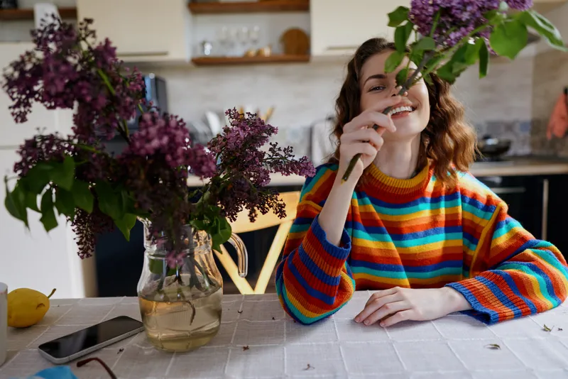 A cheerful woman sits at a home dining table, admiring a bouquet of lilac in a glass vase. Bright kitchen light, casual style, and warm atmosphere capture a simple moment of everyday happiness.