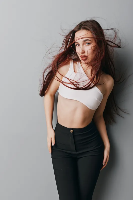 Young woman with long hair posing in white crop top and black pants against gray background. Fashion, beauty, casual style and confident expression with natural makeup and movement.