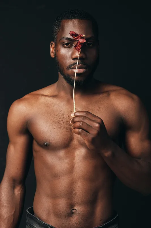 Black man with bare chest holding dried flower in front of face in creative conceptual visual style. Portrait with dark background and artistic expression of emotion and identity.