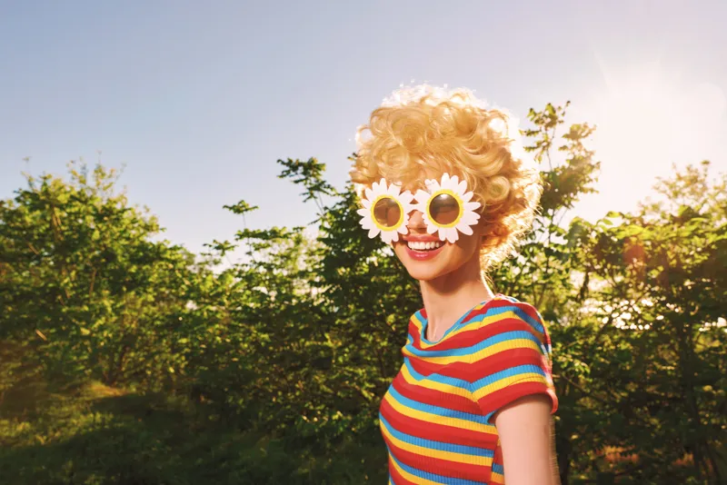 A woman in a colorful striped shirt outdoors wears playful sunglasses, smiling under bright sunlight. Retro color styling enhances a carefree lifestyle moment.