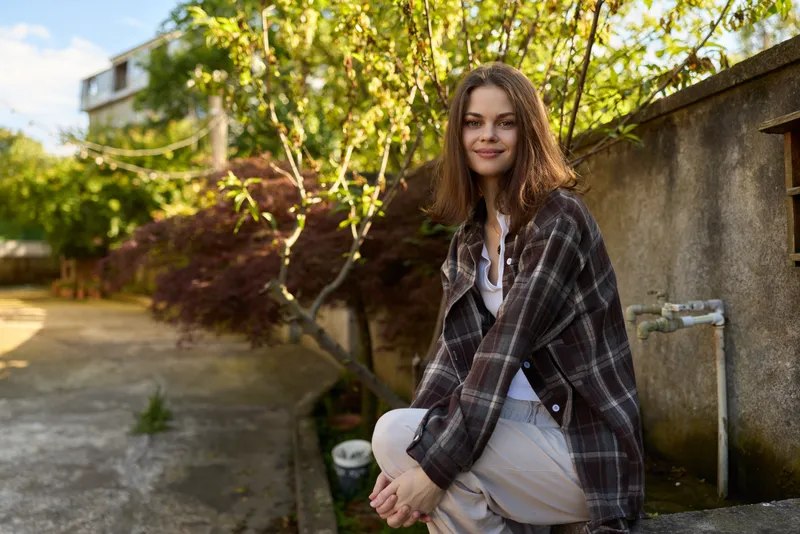 Natural woman lifestyle portrait outdoors, casual plaid jacket, smiling, relaxed pose by wall with greenery and sunlight, candid street scene under warm light