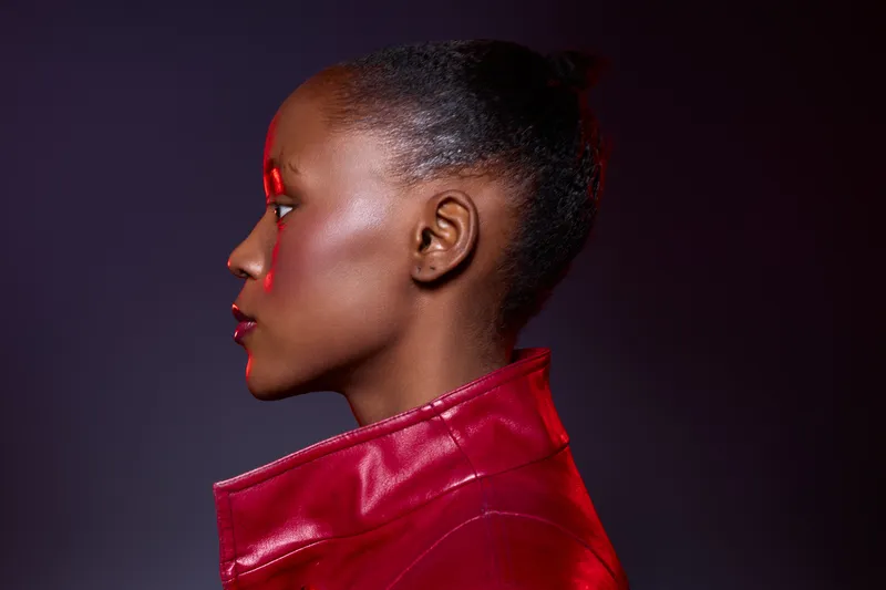 Profile of a young African woman with dark skin wearing a red leather jacket on a dark purple background. Concept of fashion, beauty, style, and confident female model portrait.