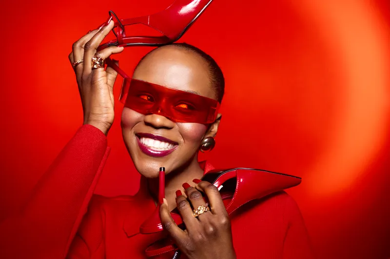 Smiling black woman with dark skin wearing red sunglasses and red outfit holds red high heels on vibrant red background. Concept of fashion, style, confidence, and beauty.