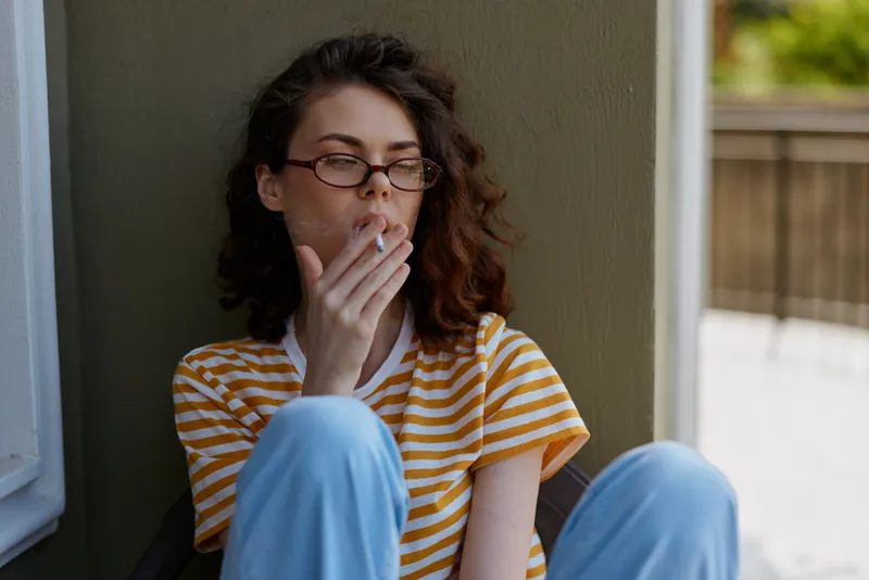 A young woman with curly hair sits against a sunlit wall on a small balcony, smoking a cigarette and looking thoughtful, wearing a striped top and light jeans in a quiet home moment.
