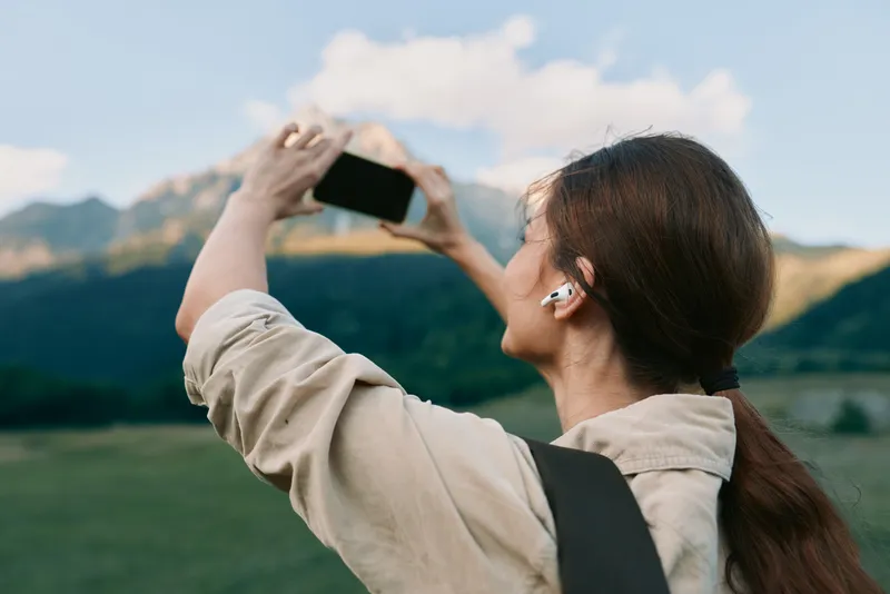 A young woman outdoors holds a phone camera to capture a photo, standing in a green field with mountains in the background, wearing a beige jacket and backpack.