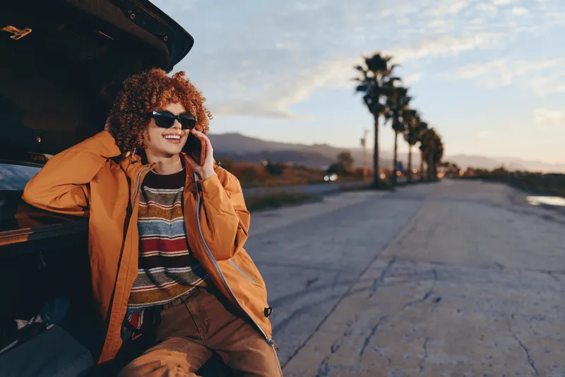 Woman lifestyle traveler in orange jacket and sunglasses sits by open car, holding smartphone near her ear, smiling at sunset roadside, casual travel moment outdoors, relaxed in distance