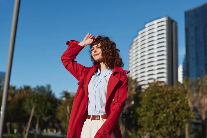 Young woman with curly hair shields her eyes in the sun, wearing a red coat and light blue shirt, standing in an urban park with tall buildings behind.
