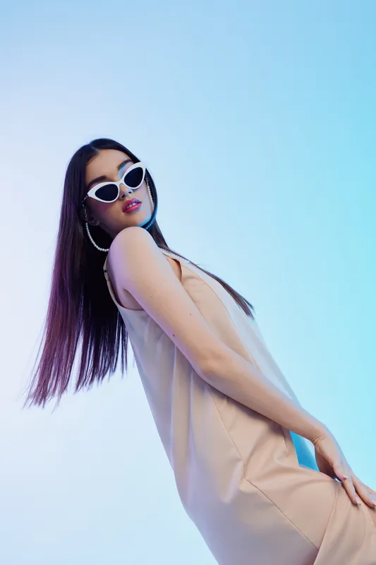 Fashionable woman with long hair and white sunglasses poses confidently in a beige dress on a blue gradient background. Concept of beauty, style, and fashion.