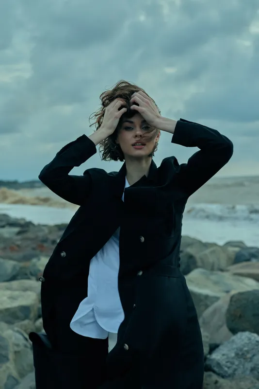 Fashion model on a rocky coastline wearing a long black coat and white shirt, wind tousling hair as she adjusts it, creating a moody, dramatic oceanfront look with dramatic clouds overhead and rugged