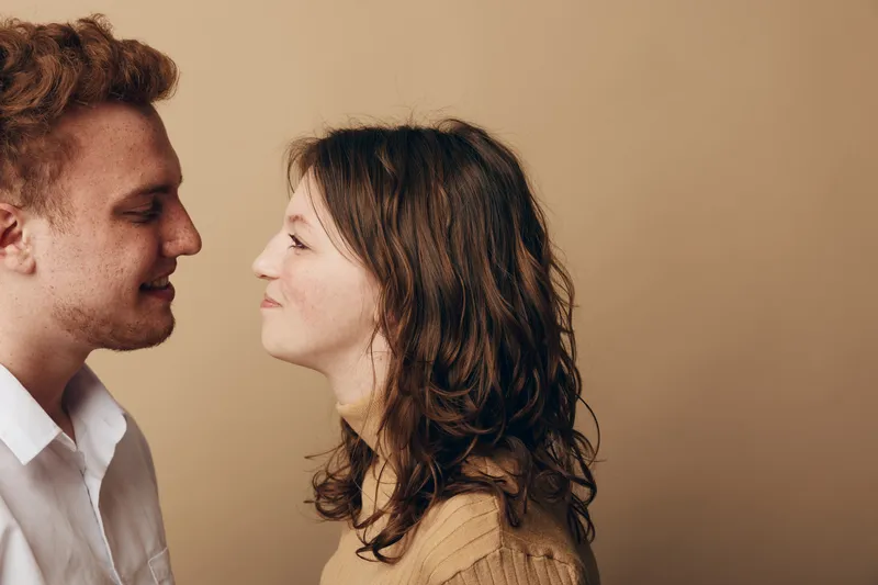 Young couple facing each other with gentle smiles, close interaction in casual clothing. Intimate moment captured on neutral background, concept of love, connection, and relationship.