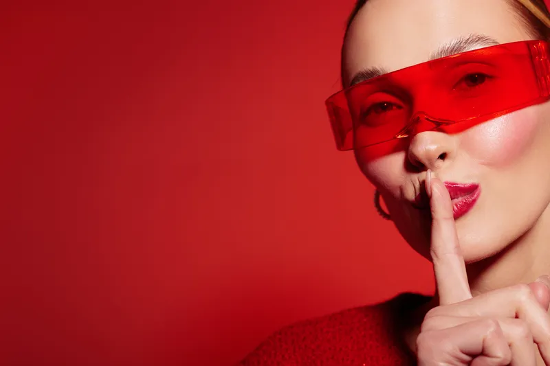 Close-up of a woman wearing red transparent glasses and red lipstick making a shushing gesture with finger on lips. Concept of secrecy, silence, and mystery on red background.