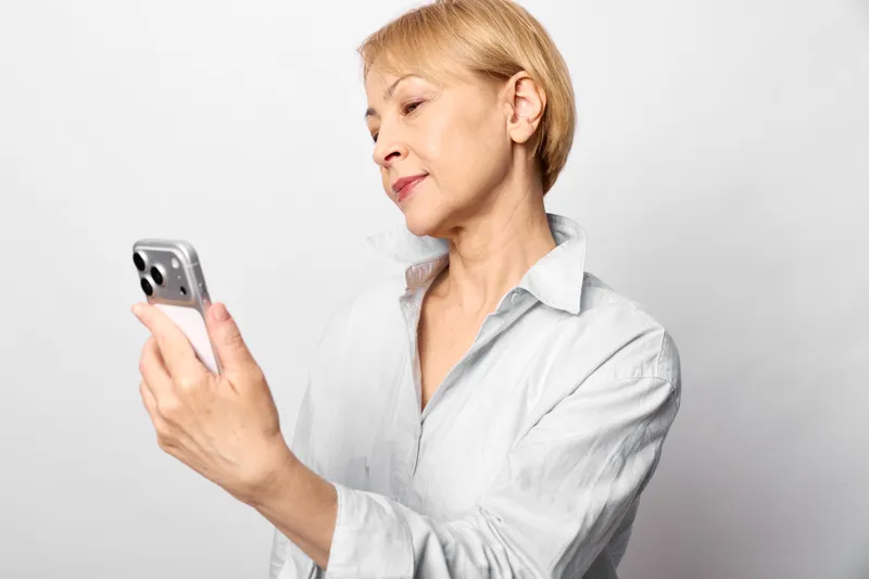 Senior woman in casual shirt holding smartphone in her hands, reading the screen with calm thoughtful expression, studio portrait on clean white background
