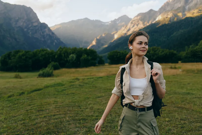 Cheerful woman outdoors, walking through a sunlit meadow with mountains in the distance, casual hiking gear, backpack, relaxed expression, natural colors, fresh breeze, serene mood