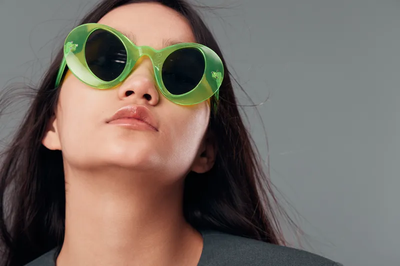 Woman with long dark hair wearing large green sunglasses on gray background. Closeup portrait with confident expression and minimal makeup in casual outfit for fashion.