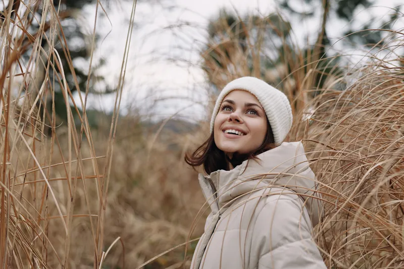 Woman outdoors in a warm coat and hat among grass, smiling as she gazes upward, capturing a serene autumn moment in natural scenery for lifestyle use.