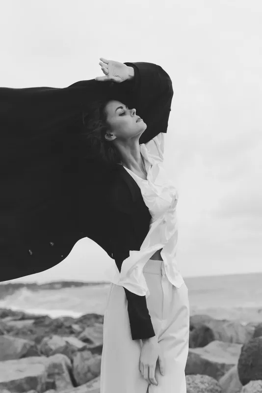 Fashion, wind and flowing coat grace a windswept beach with a distant sea, a monochrome scene that captures movement and elegance in an outdoor artistic stock image