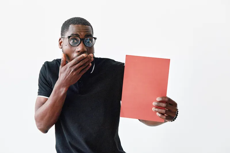 African American man with dark skin and glasses holding red folder with surprised expression on white background. Casual clothing and emotional reaction concept.