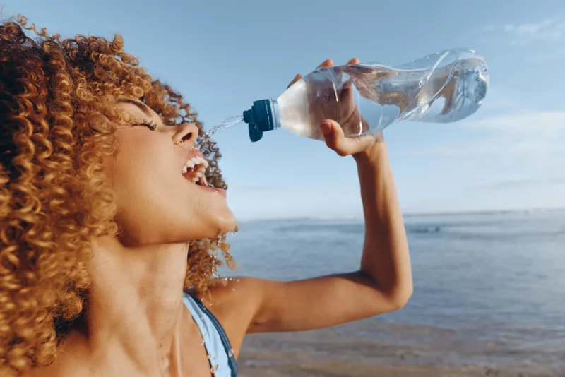 Young woman with curly hair drinks water from a clear bottle by the beach on a sunny day, enjoying a refreshing moment with the ocean and sky in the background.