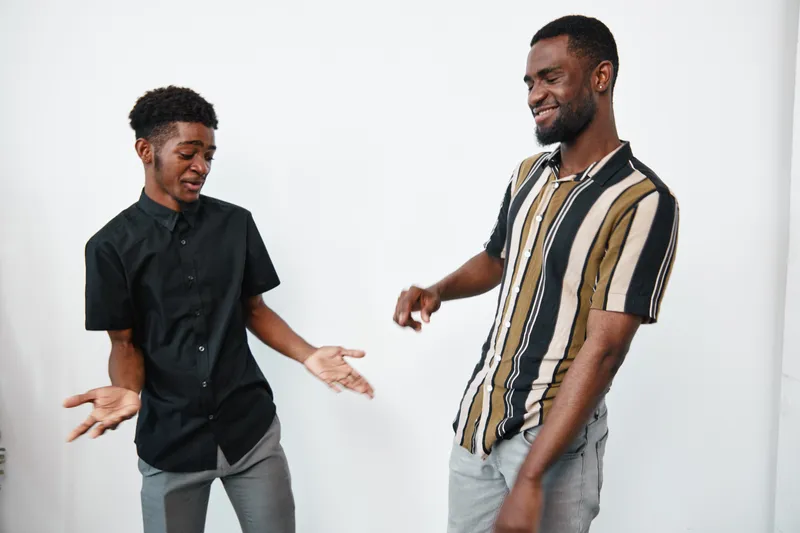 Two young black men with dark skin smiling and dancing together in casual clothes against white background in studio. Friendship and joy with happy emotions and movement.