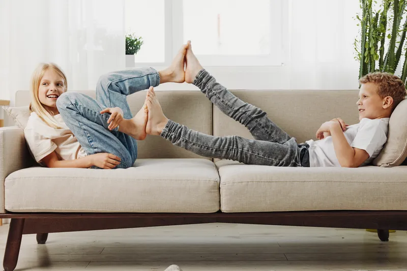 Happy children playing together on beige sofa at home, touching feet and laughing. Bright living room with natural light and plants. Concept of childhood fun and family bonding.