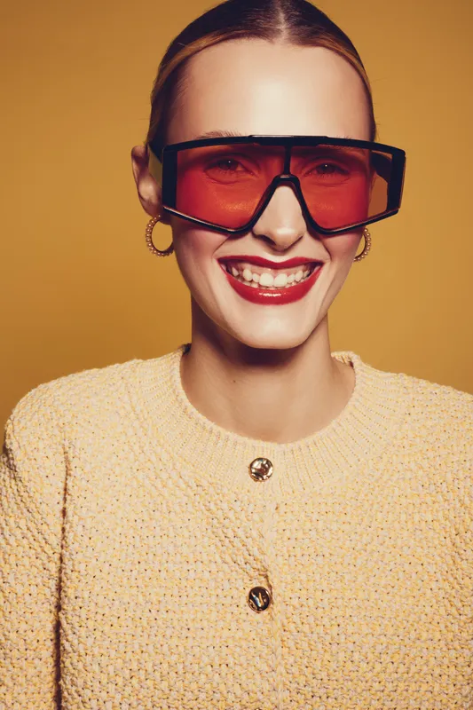 Smiling woman with red sunglasses and red lipstick wears yellow textured jacket. Studio portrait on yellow background. Concept of fashion, style, beauty, and confident mood.