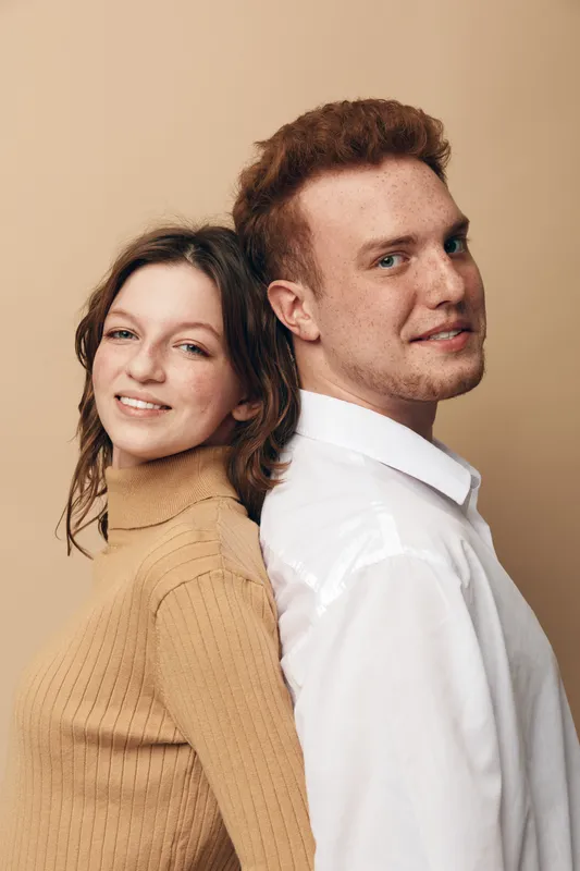 Smiling young man and woman stand back to back against beige background. Casual clothing, natural expressions, and warm lighting create friendly and relaxed atmosphere.