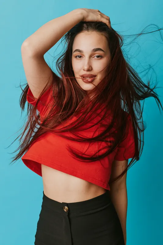 Young woman with long brown hair wearing red crop top and black pants posing with hand on head against blue background. Confident model with natural makeup and casual style.