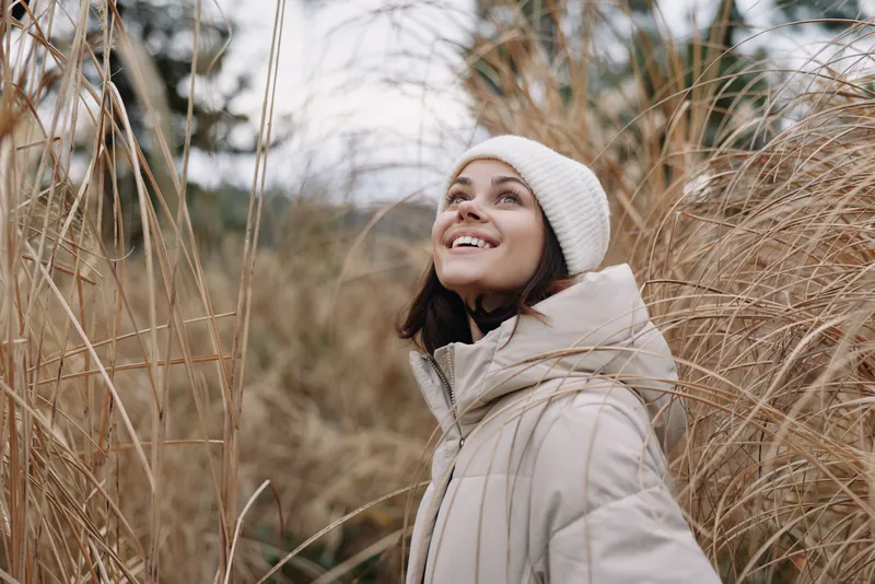 Woman in a warm coat and beanie outdoors among tall grasses, smiling and looking up at the sky during a chilly nature moment, candid scene