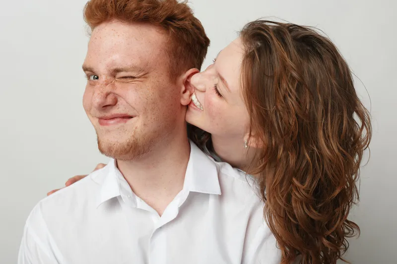Happy young woman with curly hair kisses smiling redhead man on cheek. Close-up on white background. Concept of love, affection, friendship, and joyful relationship between couple.