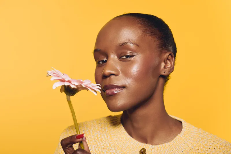 Close-up of a happy African woman with dark skin holding a pink flower near her face against yellow background. Concept of natural beauty, skincare, wellness, and self-care with floral elements.
