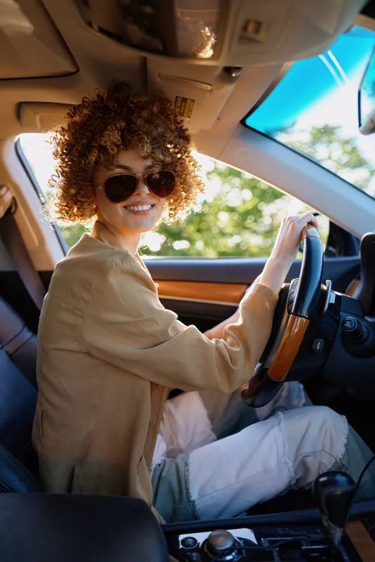 A young woman with curly hair drives a car in daylight, wearing a beige sweater and sunglasses, smiling at the wheel in a casual lifestyle moment.