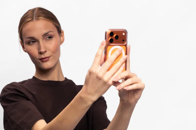 Young woman in brown shirt takes selfie with smartphone on white background. Concept of digital communication, social media, and modern technology usage by attractive female.
