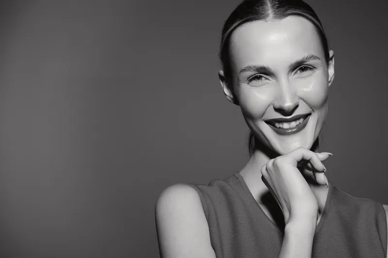 Smiling white woman with fair skin and slicked hair poses with hand near chin in black and white studio portrait. Concept for beauty and confidence.