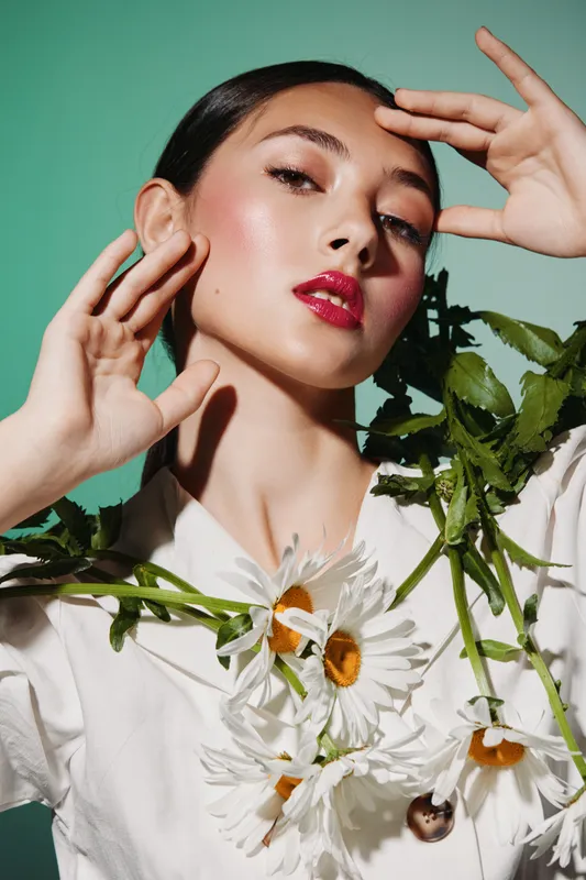 Young woman with glowing skin and bright pink lips poses with elegance holding daisies near face on a green background. Creative beauty and fashion concept highlighting vibrant makeup.