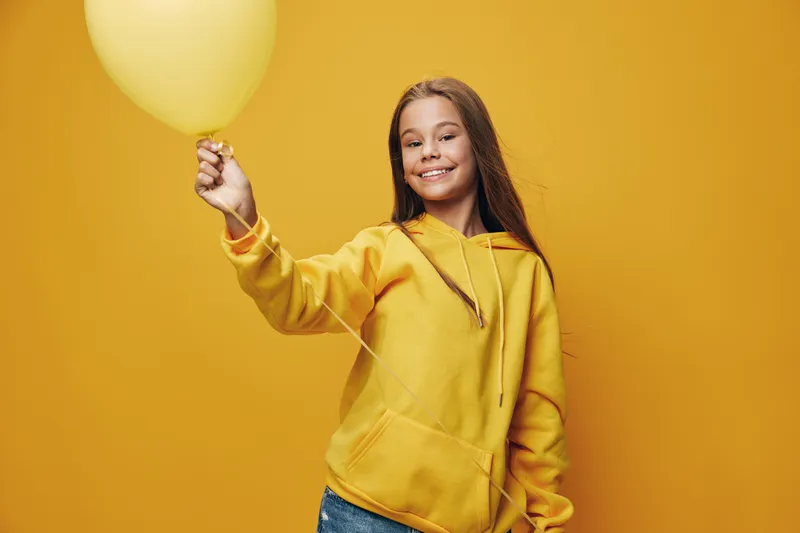 Girl in yellow hoodie smiles holding balloon against yellow background studio portrait with casual jeans happy child and colorful party decoration in minimalistic style.