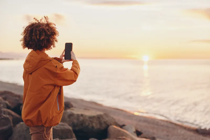 Woman lifestyle at seaside sunset, curly haired woman in orange jacket holds smartphone to take a photo on rocky shore, warm golden light, calm ocean and horizon, travel moment