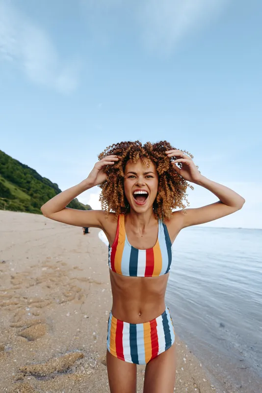 Happy young woman on a beach by the sea in a striped swimsuit, curly wet hair held with both hands, smiling toward camera in bright sunlight, scenic coastline, sand and summer vacation