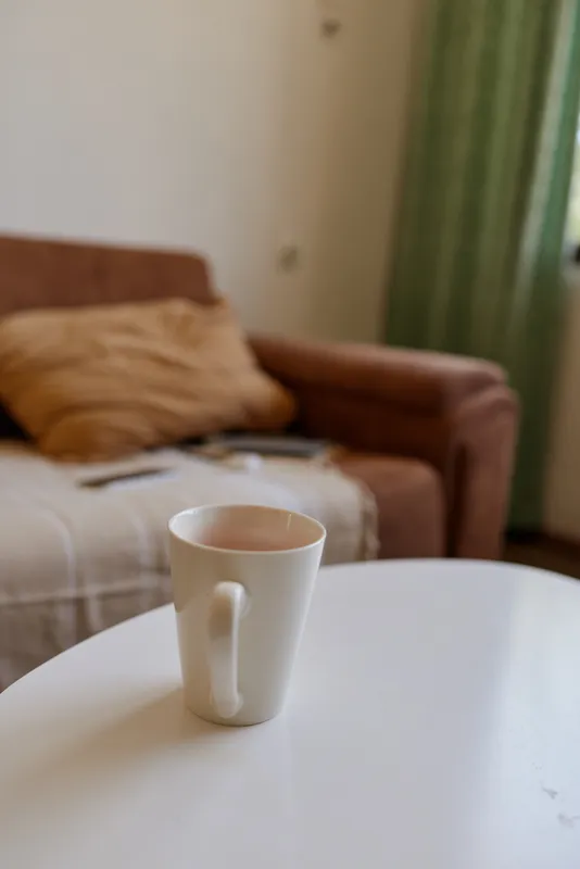 A white ceramic mug sits on a round table in a cozy living room, with a sofa and blanket in the background, conveying calm, comfort, and morning routine.