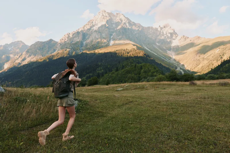 Woman hiking through a grassy meadow toward rugged mountains, carrying a backpack, enjoying fresh air and wide landscapes in a nature adventure scene.