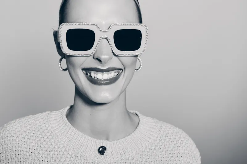 Happy white woman with light skin wearing large sunglasses and hoop earrings smiles showing teeth. Studio portrait with knitted sweater on neutral background.