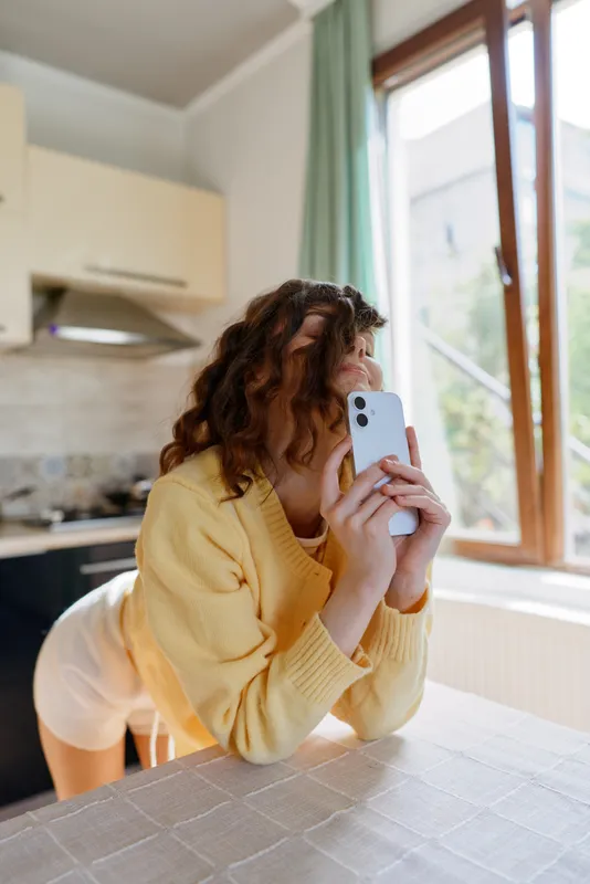 Young woman with curly hair leans on a kitchen counter wearing a soft yellow knit sweater, and takes a casual selfie with a sleek smartphone while warm daylight streams through a window.