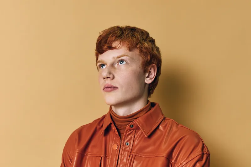 Young man with red hair wearing orange leather jacket and turtleneck looks thoughtfully to side on beige background in studio portrait with soft lighting and clear skin.